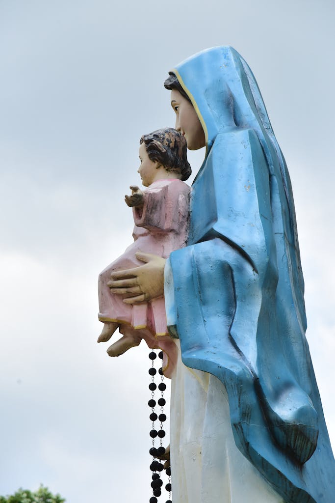 Side view of a religious statue depicting Mary holding a child with a rosary outdoors.
